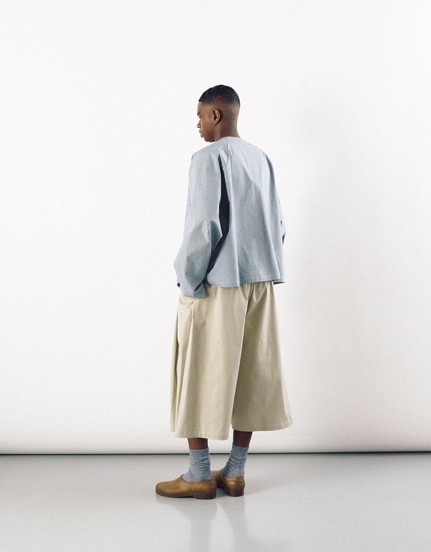 Model wearing beige wide straight-leg long shorts paired with a loose light gray top and brown shoes, showcasing minimalist unisex design with understated elegance.