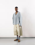 Man wearing wide straight leg beige long shorts paired with loose light blue long-sleeve top, gray socks, and brown slip-on shoes against a plain background.